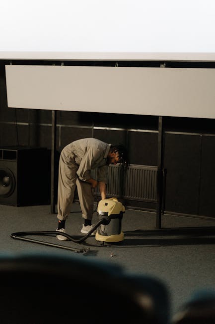 A person dressed in full protective clothing, including a white coverall suit, gloves, face mask, and disposable shoe covers, is performing deep carpet cleaning in a bright, modern living room. The in
