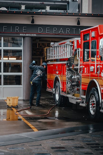 A firefighter in navy uniform is using a hose to spray water or a cleaning solution on the exterior glass and metal surfaces of the fire department building near Colliers Wood station. The building fe
